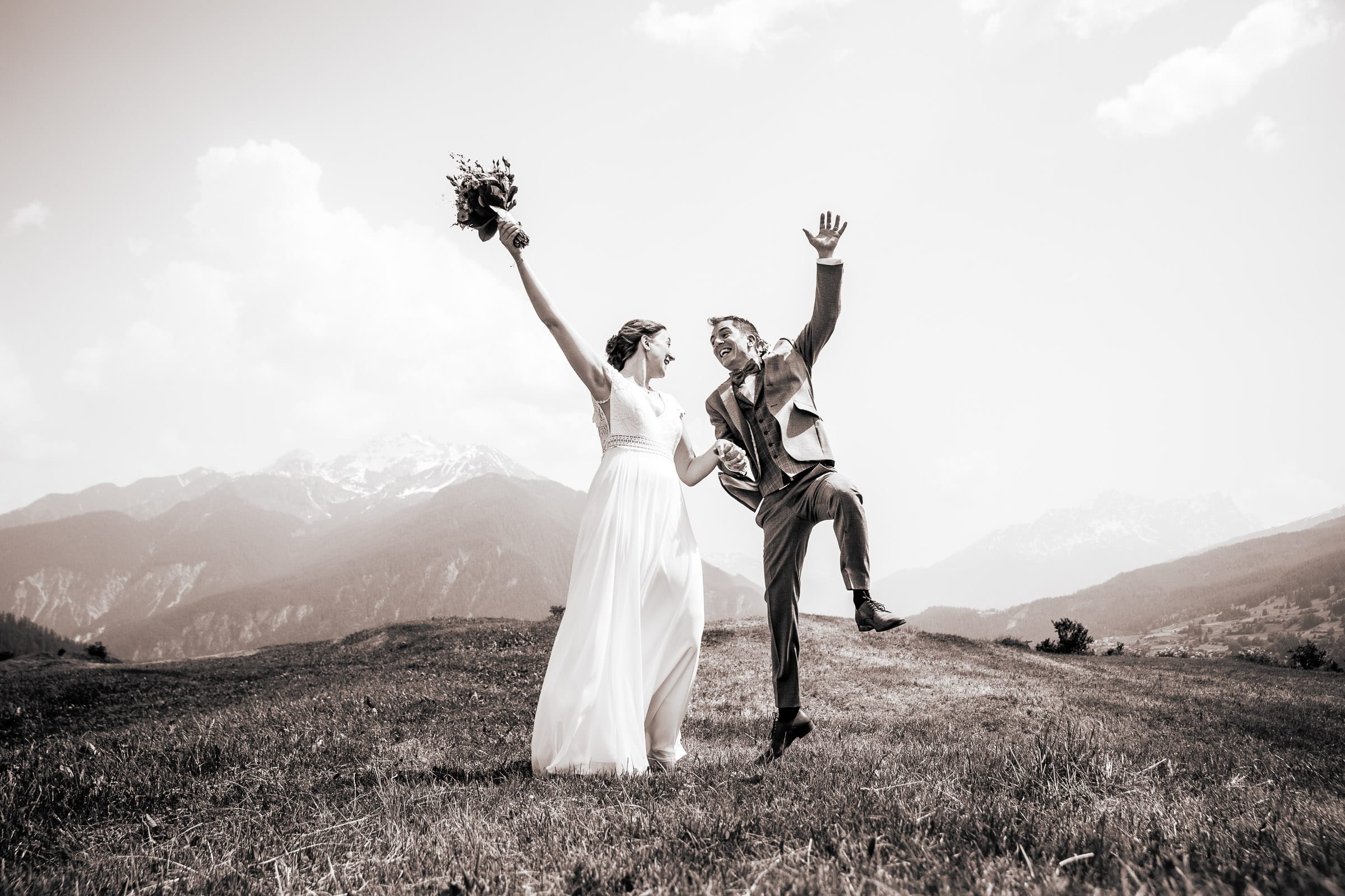 Ein Brautpaar springt freudestrahlend auf einer Bergwiese in Lenzerheide, Graubünden, fotografiert von Adrian Flütsch von Traumhochzeitsbilder. Im Hintergrund erheben sich majestätische Alpen unter einem klaren Himmel.